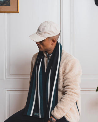 Man sitting in front of a white wall, wearing a college merino wool scarf, a beige fleece jacket, and a beige hat